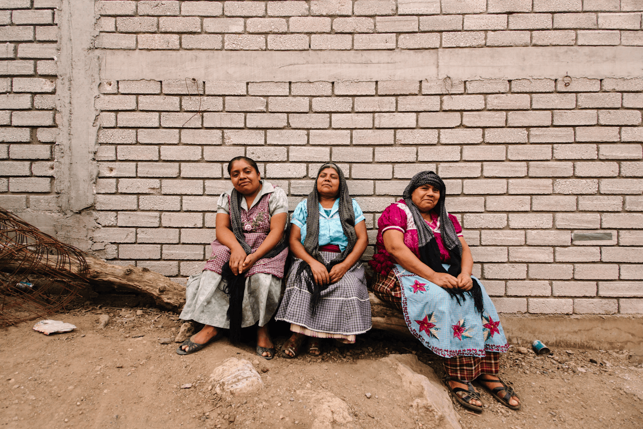 Three Mexican women in traditional clothing sit by a rustic brick wall, embodying rural culture.