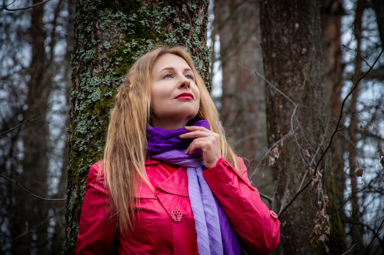 A woman in a red coat and purple scarf poses thoughtfully in a forest.